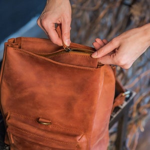 May include: A cognac-colored leather backpack featuring a main zippered compartment and a smaller front pocket with a brass-toned metal ring detail. The bag has a smooth, matte finish and visible stitching along the edges of the front pocket.