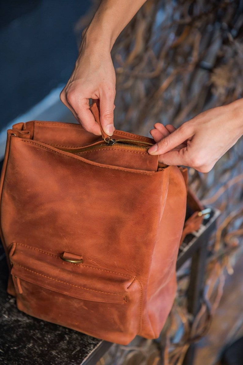 May include: A cognac-colored leather backpack featuring a main zippered compartment and a smaller front pocket with a brass-toned metal ring detail. The bag has a smooth, matte finish and visible stitching along the edges of the front pocket.
