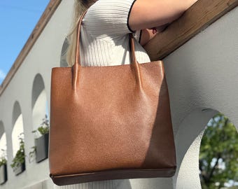 May include: A brown leather tote bag with a textured surface. The bag has two handles and a rectangular shape. The bag is being held by a person wearing sunglasses and a white top.