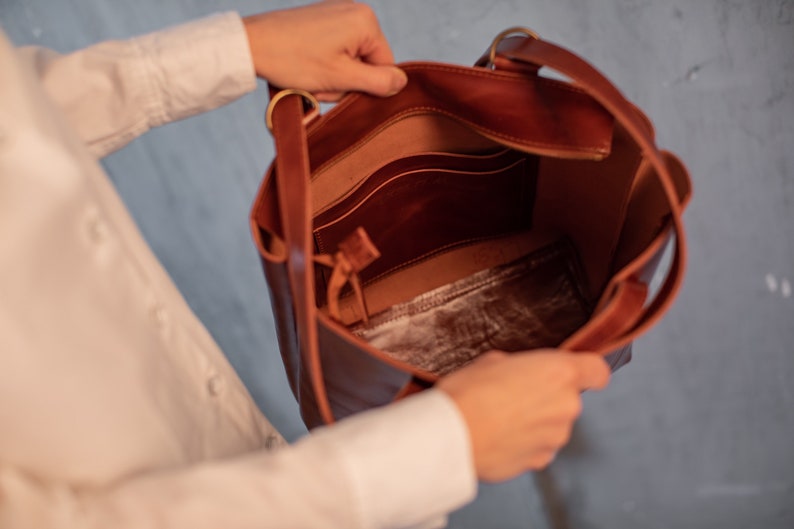 Puede incluir: Un bolso tote de cuero marr&oacute;n con un bolsillo con cremallera y un bolsillo m&aacute;s peque&ntilde;o en el interior. El bolso est&aacute; abierto y el interior es visible.