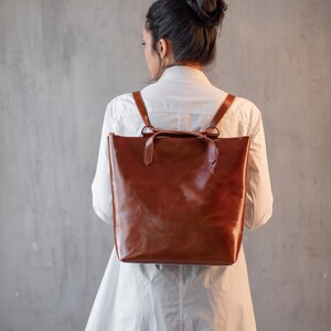May include: A brown leather backpack with adjustable straps. The backpack is worn by a person with long dark hair pulled back in a bun. The person is wearing a white shirt and black pants.