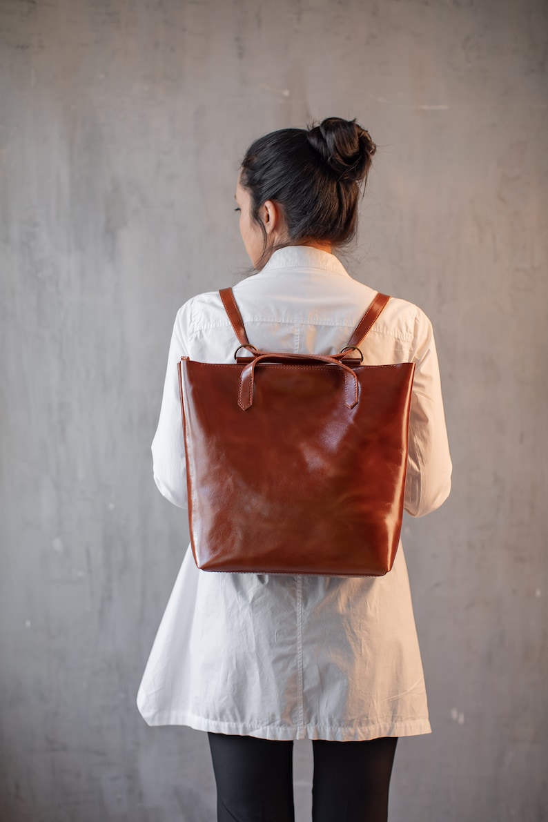 May include: A brown leather backpack with adjustable straps. The backpack is worn by a person with long dark hair pulled back in a bun. The person is wearing a white shirt and black pants.