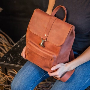 May include: A rust-colored leather backpack featuring a top flap with a metal hook closure, a front zippered pocket, and a top handle. The backpack has a matte finish and is constructed with visible stitching along the edges of the flap and body.