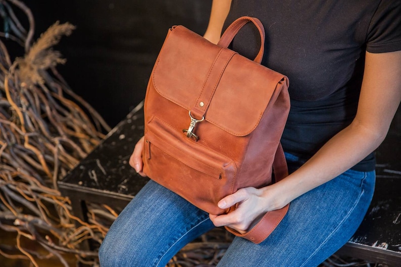 May include: A rust-colored leather backpack featuring a top flap with a metal hook closure, a front zippered pocket, and a top handle. The backpack has a matte finish and is constructed with visible stitching along the edges of the flap and body.