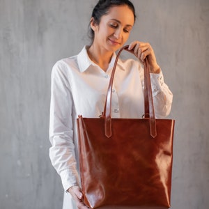 Puede incluir: Una mujer sostiene una bolsa de mano de cuero marr&oacute;n con dos asas. La bolsa tiene un acabado liso y pulido.