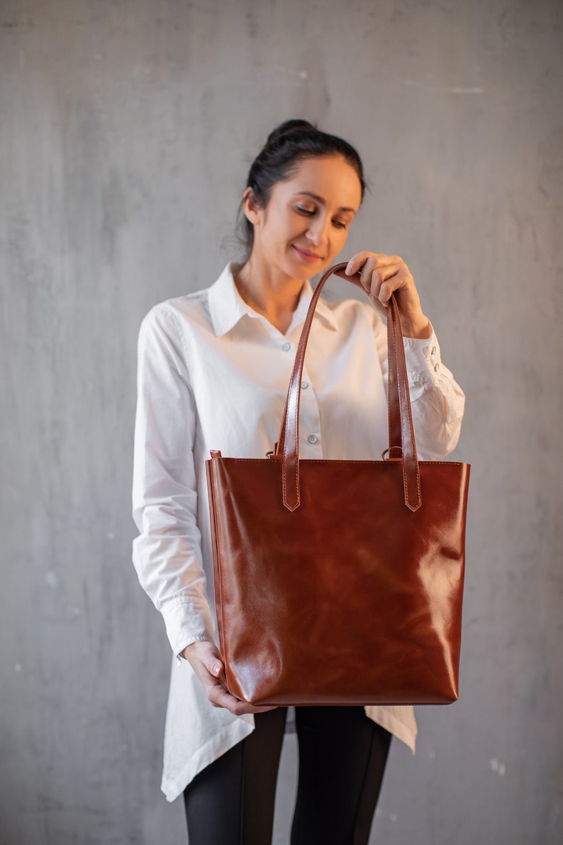 Puede incluir: Una mujer sostiene una bolsa de mano de cuero marr&oacute;n con dos asas. La bolsa tiene un acabado liso y pulido.