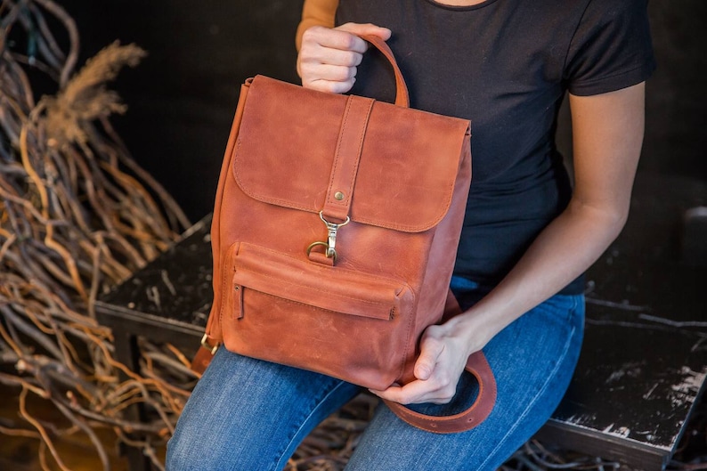May include: A rust-colored leather backpack featuring a top handle, a fold-over flap with a brass-toned metal clasp, and a front zippered pocket. The backpack has adjustable leather shoulder straps and a smooth, matte finish throughout the exterior.