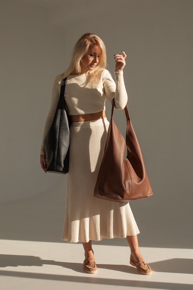 May include: A woman wearing a white crop top and a long white skirt is holding two large leather tote bags. One bag is black and the other is brown.