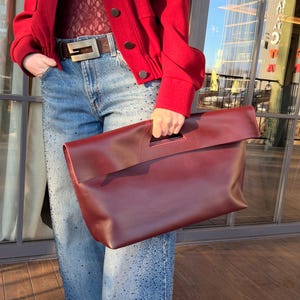 以下が含まれることがあります： 持ち手付きのバーガンディ色の革製クラッチバッグが人の前に持たれています。クラッチバッグは赤いジャケット、バーガンディ色のトップス、青いジーンズと組み合わせています。ジャケットには大きなボタンがあり、ジーンズには斑点模様があります。