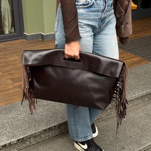 Puede incluir: Un gran bolso de mano de cuero marrón oscuro con asa y largos flecos. El bolso es rectangular y parece estar hecho de un material liso. Los flecos cuelgan del borde inferior del bolso.