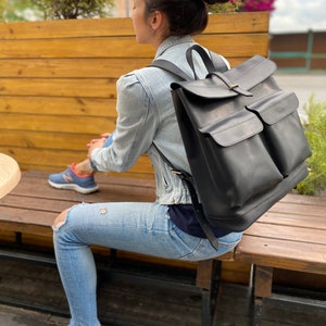 May include: A black leather backpack with two large front pockets and a flap closure. The backpack is worn by a person sitting on a wooden bench.