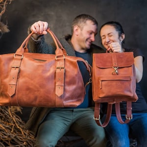 May include: A large tan leather duffel bag with two top handles and two vertical buckled straps, alongside a smaller matching tan leather backpack featuring a front zippered pocket, a fold-over flap with a metal clasp, and adjustable shoulder straps.