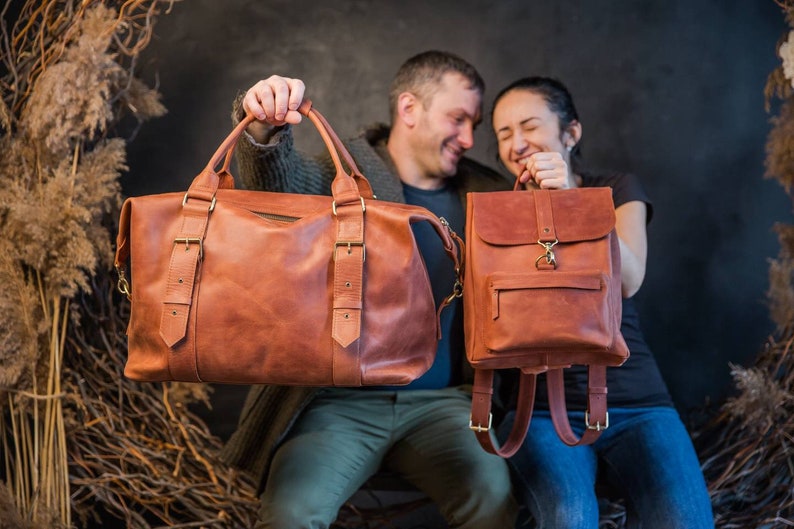 May include: A large tan leather duffel bag with two top handles and two vertical buckled straps, alongside a smaller matching tan leather backpack featuring a front zippered pocket, a fold-over flap with a metal clasp, and adjustable shoulder straps.