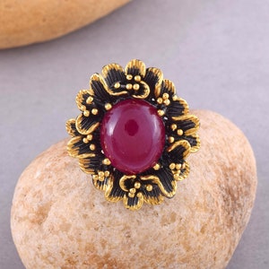 May include: A ring with a large, dark red gemstone surrounded by a black and gold floral design.