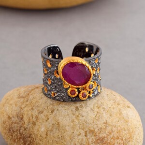 Peut inclure: Une large bague en argent avec une finition oxydée noire et une grosse pierre précieuse rose. La bague est décorée de petites pierres précieuses roses.