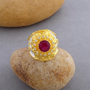 Op de afbeelding: Een gouden ring met een grote rode edelsteen omringd door kleinere witte edelstenen. De ring heeft een getextureerd ontwerp.