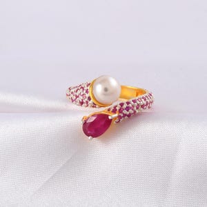 May include: A gold-toned ring featuring a large, round, white pearl and a teardrop-shaped ruby. The ring band is encrusted with small, pink gemstones. The ring is set against a white, textured background.