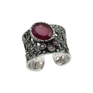 May include: A wide silver ring featuring a textured, porous surface with a large oval-shaped dark pink gemstone set in the centre. Below the main stone, three smaller round red gemstones are arranged in a cluster on the metal band.
