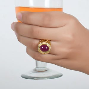 Könnte beinhalten: Ein goldener Ring mit einem großen, ovalen, dunkelroten Edelstein, der von kleineren, klaren Edelsteinen umgeben ist. Der Ring hat ein detailliertes, kompliziertes Design.