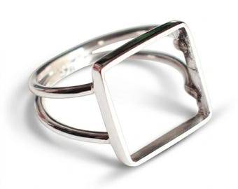 Op de afbeelding: Een zilverkleurige ring met een hol, vierkant middendeel bevestigd aan een dubbele ringband. Het metaal heeft een gladde, gepolijste afwerking en het vierkante frame is open in het midden, wat zorgt voor een geometrisch ontwerp rond de vinger.