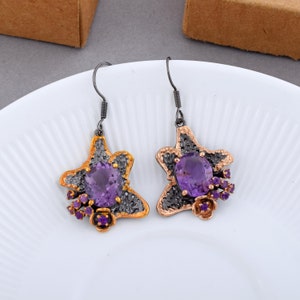 Könnte beinhalten: Ein Paar Hängeohrringe mit einem sternförmigen Design. Die Ohrringe sind aus schwarzem und goldenem Metall gefertigt und haben einen großen violetten Amethyst-Edelstein in der Mitte jedes Sterns. Die Ohrringe sind mit kleineren violetten Amethyst-Edelsteinen an den Rändern verziert.