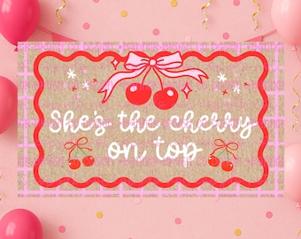 May include: A rectangular sign with the text "She's the cherry on top" in a playful font, surrounded by a red and pink border. The design includes cherries, bows, and stars, set against a textured background, with pink balloons and confetti.