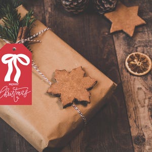 May include: A wrapped gift with a red tag that says "Merry Christmas" in white script. The gift is wrapped in brown paper and tied with twine. A gingerbread snowflake and star, pine sprigs, cinnamon stick, and dried orange slices are on a wooden surface.
