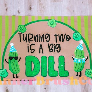May include: A rectangular banner with a brown background and green vertical stripes. The banner features the text "Turning two is a big dill" with cartoon pickle illustrations and a birthday theme.