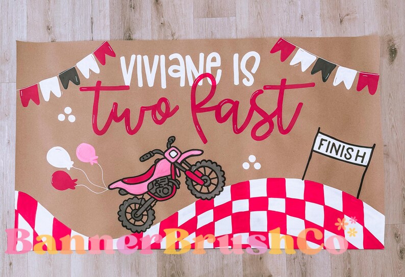 Pu&ograve; includere: Striscione marrone con la scritta "Viviane is two fast" in bianco e rosa. Lo striscione presenta una moto rosa, palloncini e un traguardo a scacchi. I gagliardetti decorativi sono in alto.