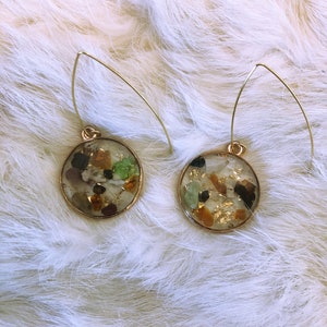 May include: A pair of gold hoop earrings with round, clear resin charms filled with small, colorful stones and gold flakes.
