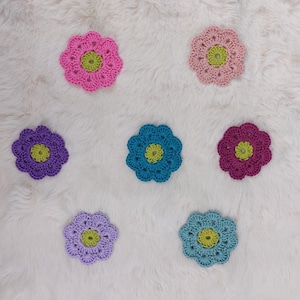 Könnte beinhalten: Sieben farbenfrohe, gehäkelte Untersetzer in Blumenform. Die Untersetzer sind in den Farbtönen Rosa, Lila, Blau und Grün gehalten. Die Mitte jeder Blume hat eine kontrastierende gelb-grüne Farbe. Die Untersetzer sind auf einer weißen, flauschigen Oberfläche angeordnet.