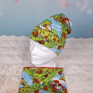 Könnte beinhalten: Ein grünes und blaues Mütze- und Halstuch-Set mit einem Cartoon-Bienen- und Mohnblumen-Design. Die Mütze befindet sich auf einem weißen Schaufensterkopf. Das Halstuch hat rote Punkte auf der Innenseite.