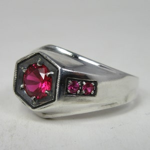 Può includere: Un anello d'argento con una grande pietra rossa sfaccettata incastonata in una lunetta esagonale. Due pietre rosse più piccole sono incastonate su entrambi i lati della pietra principale.