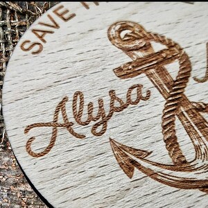 May include: A wooden round coaster with a laser-engraved anchor design. The text "Save the Date" is at the top of the coaster. The names "Alyssa & Mark" are engraved below the anchor.