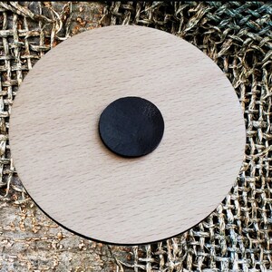 May include: A round, wooden coaster with a black circle in the center. The coaster is made of light wood and has a smooth finish.