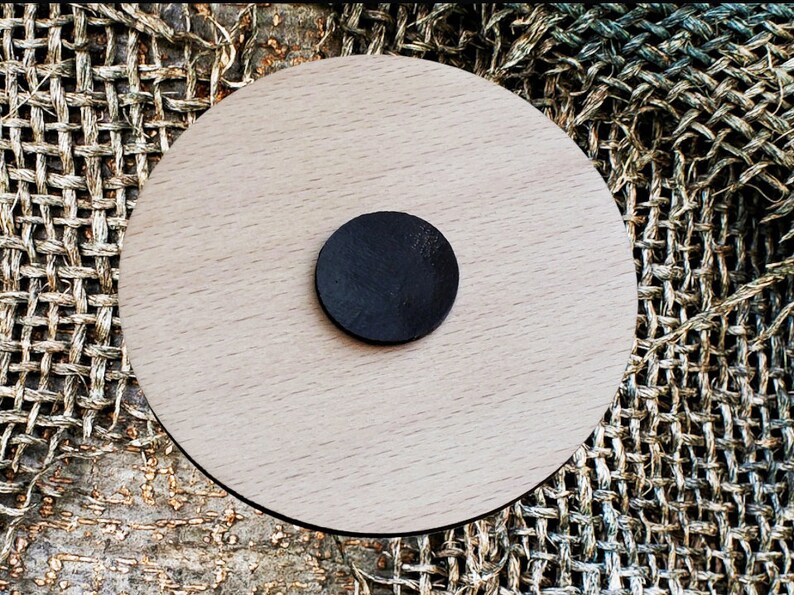 May include: A round, wooden coaster with a black circle in the center. The coaster is made of light wood and has a smooth finish.