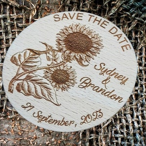 May include: A round wooden save the date sign with a sunflower design. The text reads "Save the Date" and "Sydney & Brandon" with the date "27 September, 2023".