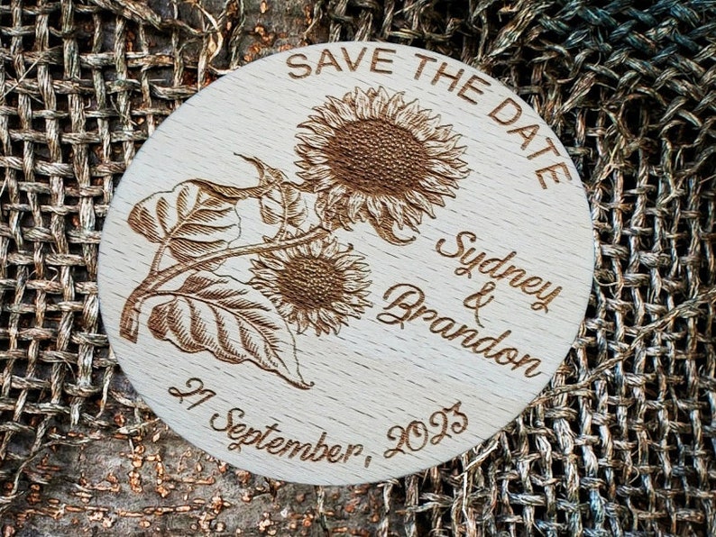 May include: A round wooden save the date sign with a sunflower design. The text reads "Save the Date" and "Sydney & Brandon" with the date "27 September, 2023".
