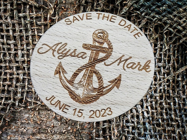 May include: A round wooden save the date sign with a nautical theme. It features a carved anchor with rope, the text "Save the Date", the names "Alyssa Mark", and the date "June 15, 2023".