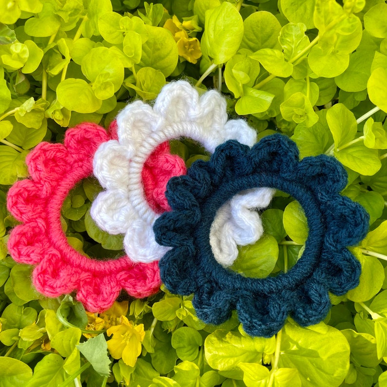 May include: Three crocheted hair scrunchies in different colors: pink, white, and dark blue. Each scrunchie has a ruffled edge.