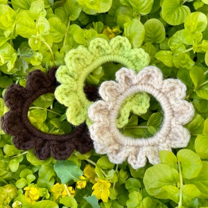 May include: Three crocheted hair scrunchies in brown, green, and beige. The scrunchies are made with a textured, ruffled design.