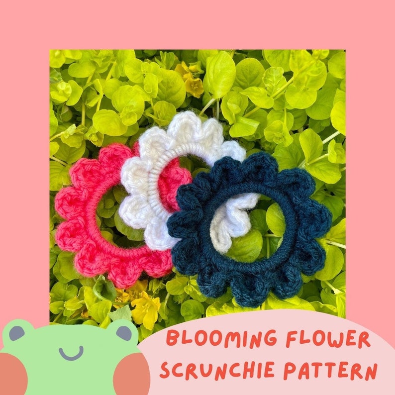 May include: Three crocheted flower-shaped scrunchies in pink, white, and dark blue. The scrunchies are arranged in a circle on a bed of green leaves.