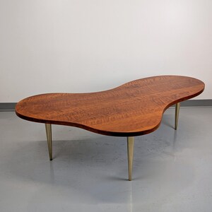 May include: A mid-century modern coffee table with a unique, free-form, organic shaped top made of wood. The table has four slender, gold-colored metal legs.