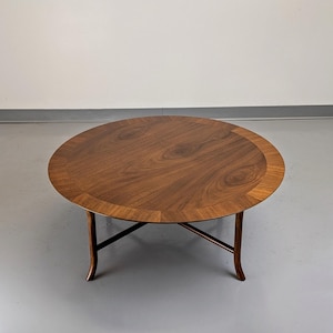 May include: Oval-shaped wooden coffee table with a dark brown finish and a lighter brown border. The table has four curved legs that meet in the center. The table is approximately 48 inches in diameter.
