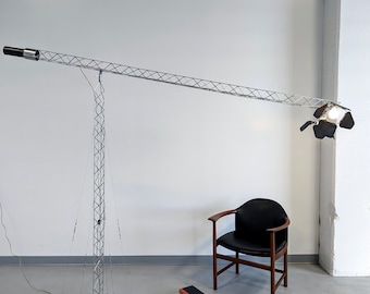 Może przedstawiać: Srebrna metalowa lampa stojąca z długim, wysuwanym ramieniem i czarnym reflektor. Lampa znajduje się w pokoju z białą ścianą i czarnym skórzanym fotelem.