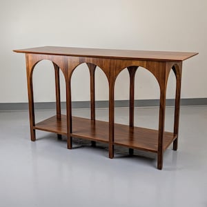 May include: A wooden console table with a rectangular top and a lower shelf. The table features arched supports and is made of a rich, dark brown wood. The design is mid-century modern.