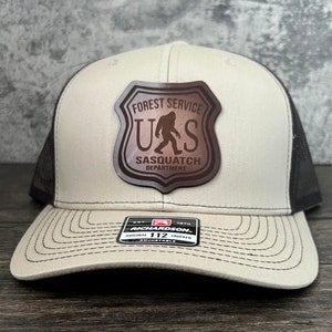 May include: A beige and black trucker hat with a brown leather patch featuring a silhouette of Bigfoot and the text "Forest Service U.S. Sasquatch Department".