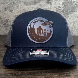 May include: A navy blue and grey trucker hat with a brown leather patch featuring a silhouette of Bigfoot and an alien with a UFO in the background. The patch is circular and has a starry night sky design.