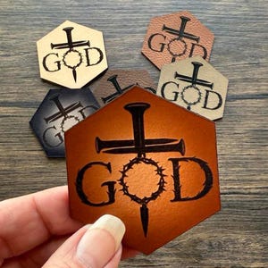 May include: Hexagon-shaped leather patches with the word "GOD" and a cross, nail, and crown of thorns design. The patches are in various colors, including brown, black, and tan. The patches are shown on a wooden surface.