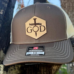 Puede incluir: Gorra de camionero marrón y beige con visera cosida. La gorra presenta un parche de cuero con la palabra "GOD" y un diseño de cruz y lanza. La gorra tiene una etiqueta Richardson 112.